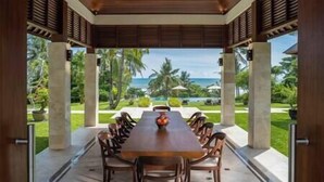 Uma área de refeições coberta com uma mesa comprida e cadeiras, com vista para uma paisagem tropical e o oceano.