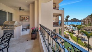 A balcony with a grill, potted plants, and a view of a resort pool and mountains. - Newly Renovated Luxury Villa! Medano Beachfront! Overlooking Pools, and Sea of Cortez! (Cabo San Lucas)