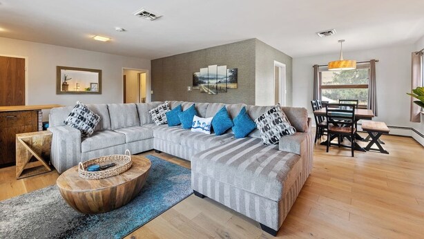 Un salon moderne avec un canapé d’angle gris, une table basse en bois et un coin repas comprenant une table et des chaises en bois.