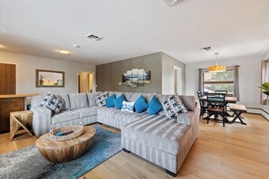 Un salon moderne avec un canapé d’angle gris, une table basse en bois et un coin repas comprenant une table et des chaises en bois.