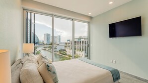 A bedroom with a large window, a bed, a TV, and a view of a cityscape. - Modern apartment in Puerto Cancun with partial ocean and marina view 🌊 (Puerto Cancun)