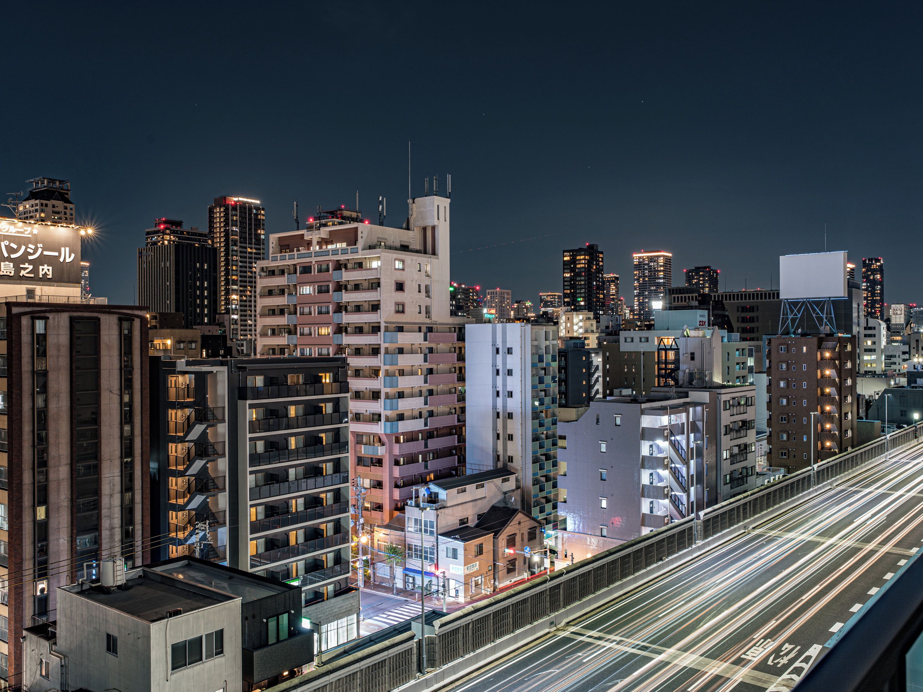 A cityscape at night with illuminated buildings and a long exposure of light trails on the road.
