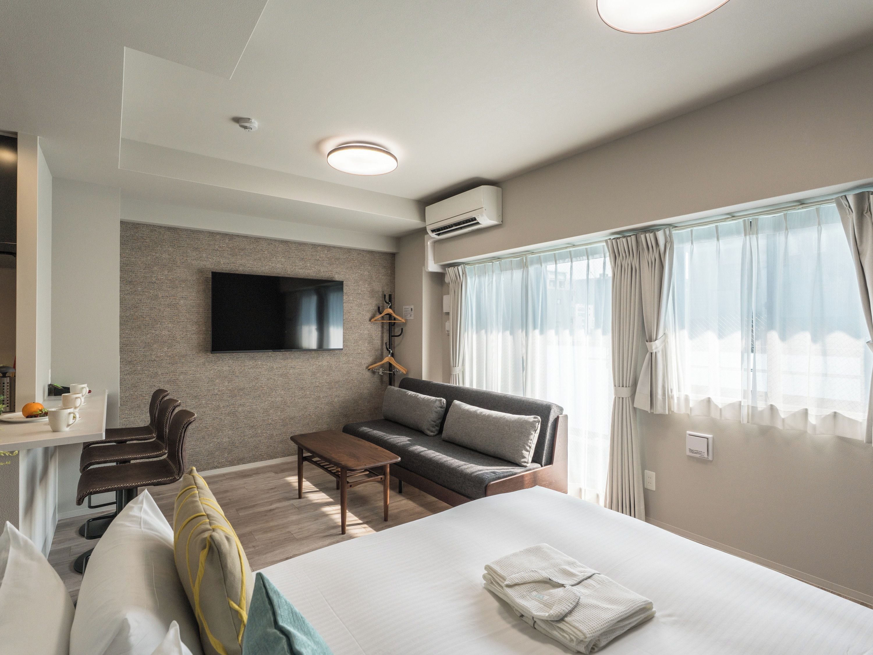 A hotel room with a bed, a sofa, a TV mounted on the wall, a desk with a chair, and a window with curtains.