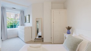 A bedroom with a bed, a dresser, a mirror, a wardrobe, and a window with curtains. - GuestReady - Charming Retreat in Vibrant London (London)