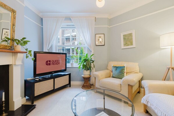 A living room with a television displaying 'GUEST READY,' a sofa, a chair, a glass coffee table, and a fireplace. - GuestReady - Charming Retreat in Vibrant London (London)