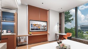 A modern hotel room with a large bed, a TV mounted on the wall, a small desk with a computer, a window with a city view, and a small table with a glass of wine and flowers. - From·K Hotel Pazhou (Guangzhou)