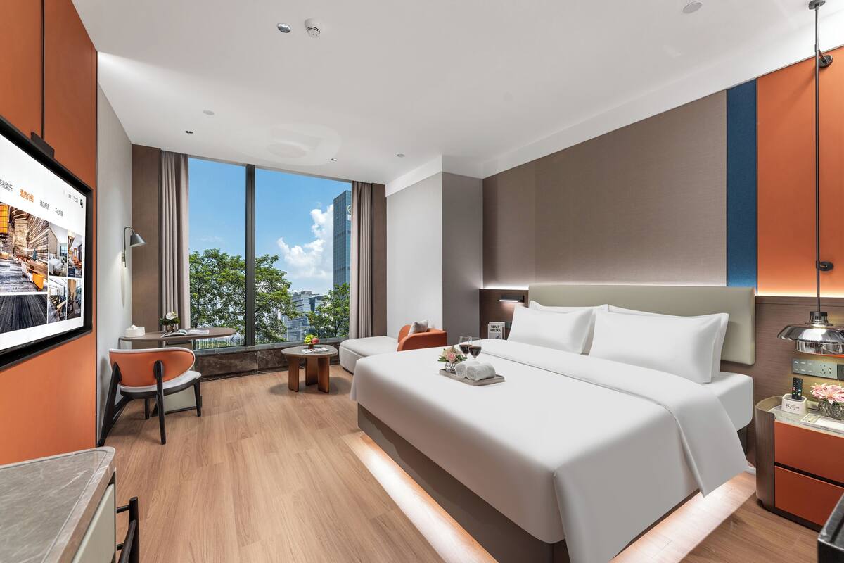 Una habitación de hotel moderna con una cama grande, un escritorio, una silla y una mesita pequeña. Cuenta con un ventanal amplio que ofrece vistas a la ciudad.