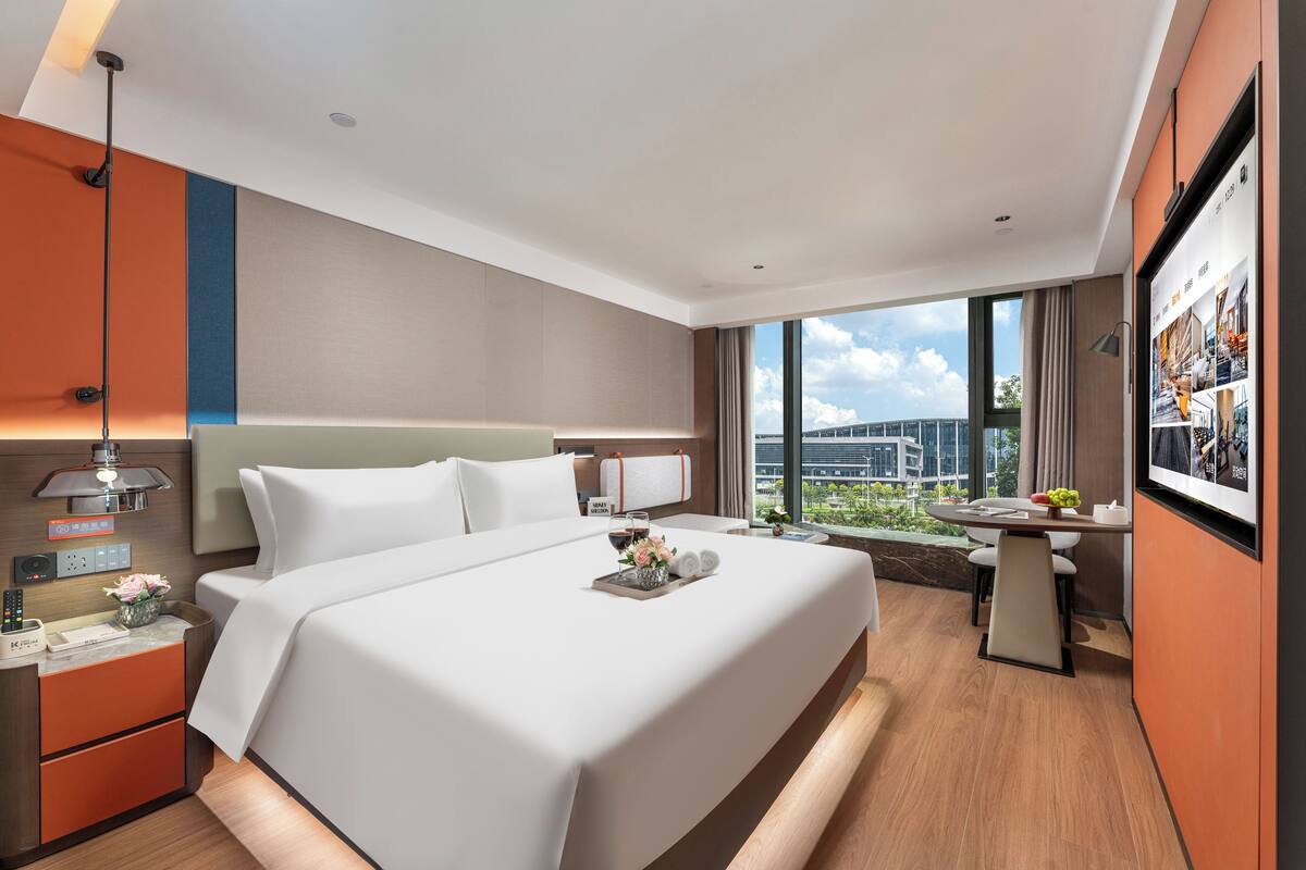 Una habitación de hotel moderna con una cama grande, mesita de noche y un escritorio con silla. La habitación tiene un ventanal amplio con vistas a la ciudad.