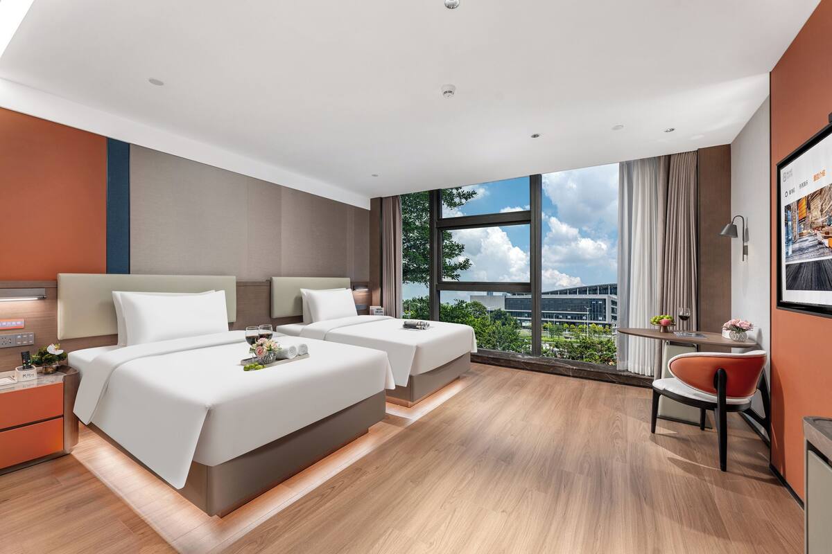 Una habitación de hotel moderna con dos camas, una mesita con una silla y un ventanal que ofrece vistas a la ciudad.