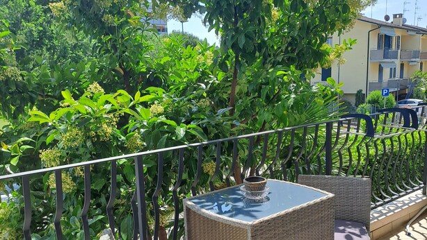 A balcony with wicker furniture and a glass table, surrounded by lush greenery and a clear sky.