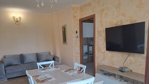 A living room with a sofa, a dining table with chairs, a TV mounted on the wall, and a bookshelf.