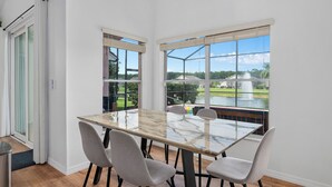 Un comedor con una mesa de vidrio y cuatro sillas, ventanas amplias con persianas y vistas a un lago y follaje.
