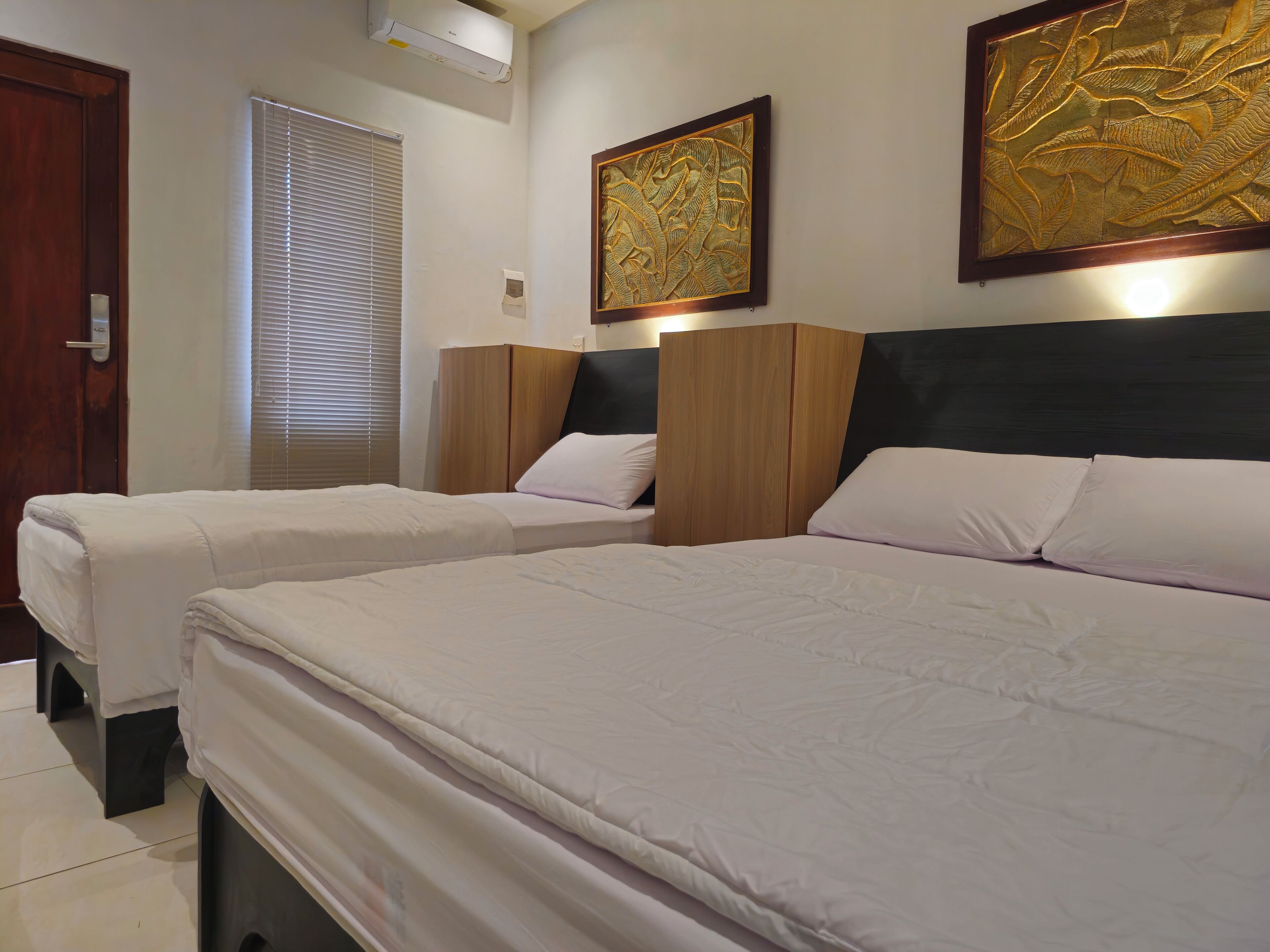 Une chambre d’hôtel avec deux lits, une armoire en bois, un climatiseur fixé au mur et une fenêtre avec des stores.