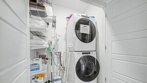 Une buanderie équipée de deux machines à laver et deux sèche-linge superposés, d’un balai, de produits de nettoyage et d’un panneau indiquant qu’il faut nettoyer le filtre à peluches avant et après chaque utilisation.