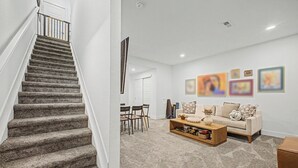 Un escalier recouvert de moquette mène à une porte blanche, puis on découvre un salon avec un canapé beige, une table basse et un coin repas avec des chaises.