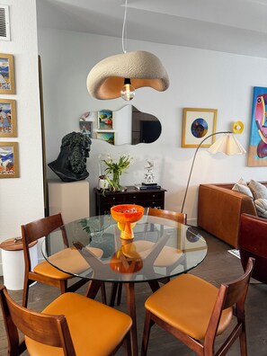 Un espace repas moderne avec une table en verre, des chaises en bois, un luminaire suspendu original et diverses œuvres d’art accrochées aux murs.