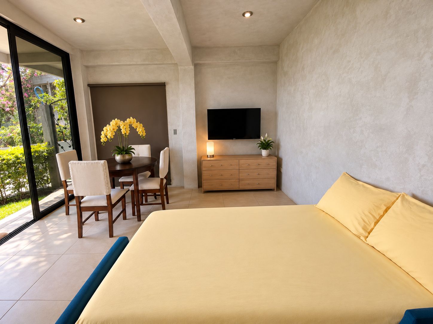 Un dormitorio moderno con una cama grande, televisión, zona de comedor con sillas y vistas a través de un ventanal.