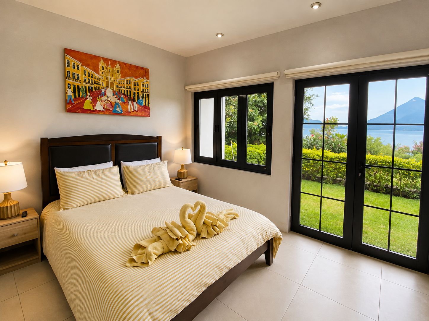 A bedroom with a large bed, two bedside tables, a painting, and a view of a mountain and lake through large windows.