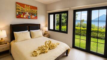 A bedroom with a large bed, two bedside tables, a painting, and a view of a mountain and lake through large windows.