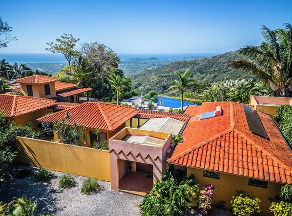 A resort with multiple buildings featuring red-tiled roofs, surrounded by lush greenery and a view of the ocean.