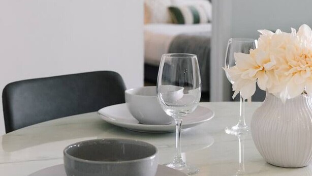 A dining area with a table set for two, a vase with flowers, and a bed in the background.
