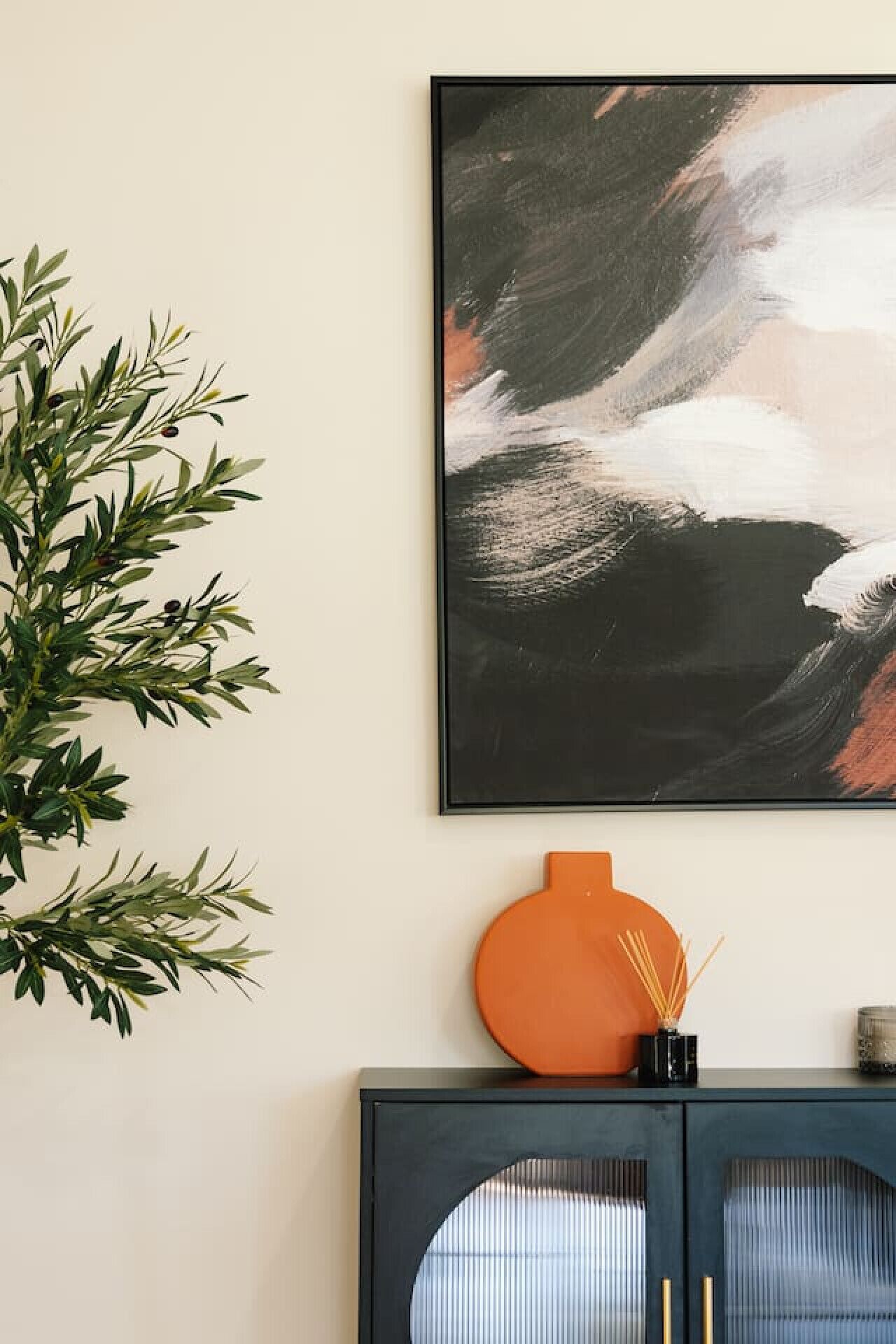 A framed abstract painting on a wall, a potted plant, and a cabinet with a vase and incense sticks.