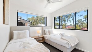 Habitación de hotel con dos camas, ventilador de techo y ventanas amplias con vistas a árboles y edificios.