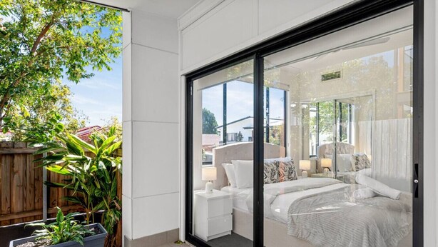Un dormitorio con un gran ventanal corredera, una cama con ropa de cama blanca, una mesita de noche y una planta en maceta.