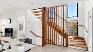 Un interior moderno con una escalera de madera, una zona de comedor con una mesa y sillas blancas, y un televisor en la pared.