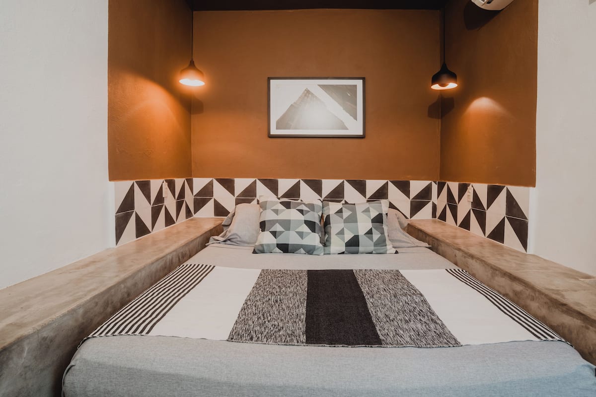 Ein Zimmer mit einem Bett, einem geometrisch gemusterten Kopfteil, zwei Nachttischen, einem gerahmten Bild an der Wand und zwei Wandleuchten.