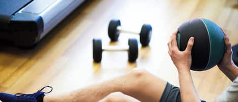 A person sitting on a mat, holding a medicine ball, with a treadmill and dumbbells in the background.