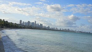A beach with waves lapping at the shore, a city skyline in the distance, and a clear sky with clouds. - CABO BRANCO BEACHFRONT FLAT at the Green Sunset Hotel (João Pessoa)