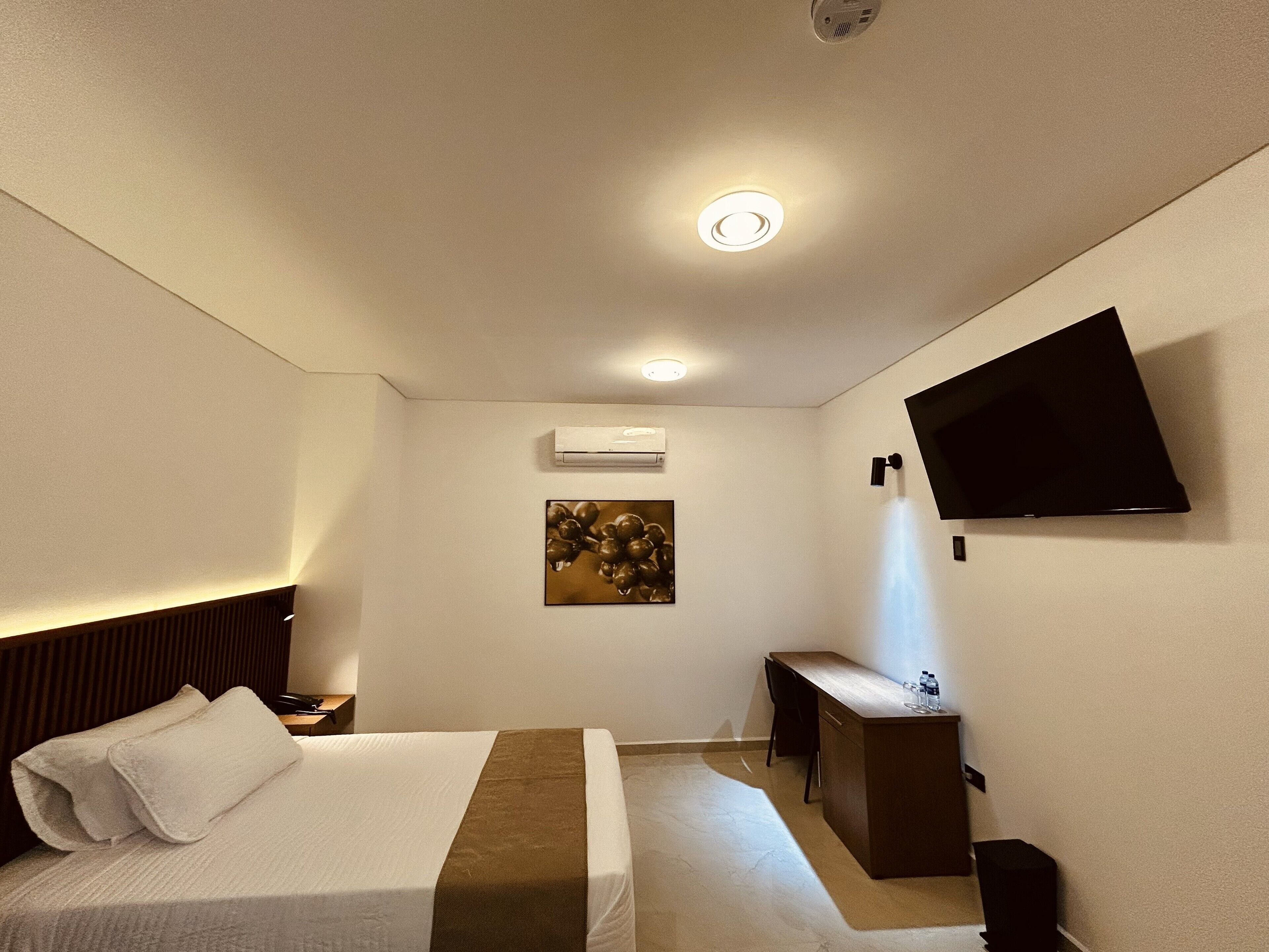 A hotel room with a bed, bedside table, desk, chair, flat-screen TV, artwork, and an air conditioning unit.