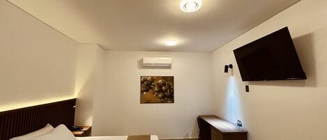 A hotel room with a bed, bedside table, desk, chair, flat-screen TV, artwork, and an air conditioning unit.