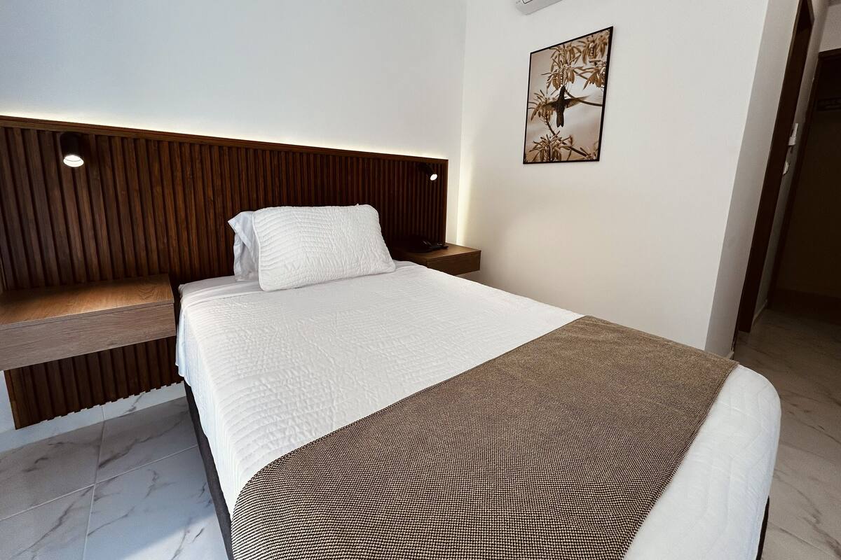 Moderni hotellihuone, jossa on sänky, puinen pääty, yöpöytä ja seinälle asennettu ilmastointilaite.