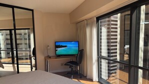 Une chambre d’hôtel avec un lit, un bureau sur lequel est posée une télévision diffusant une scène de plage, une chaise et de grandes fenêtres avec des rideaux.