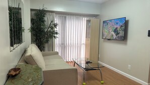 Un salon comprenant un canapé, une table basse en verre, un téléviseur fixé au mur et une grande fenêtre avec des rideaux.