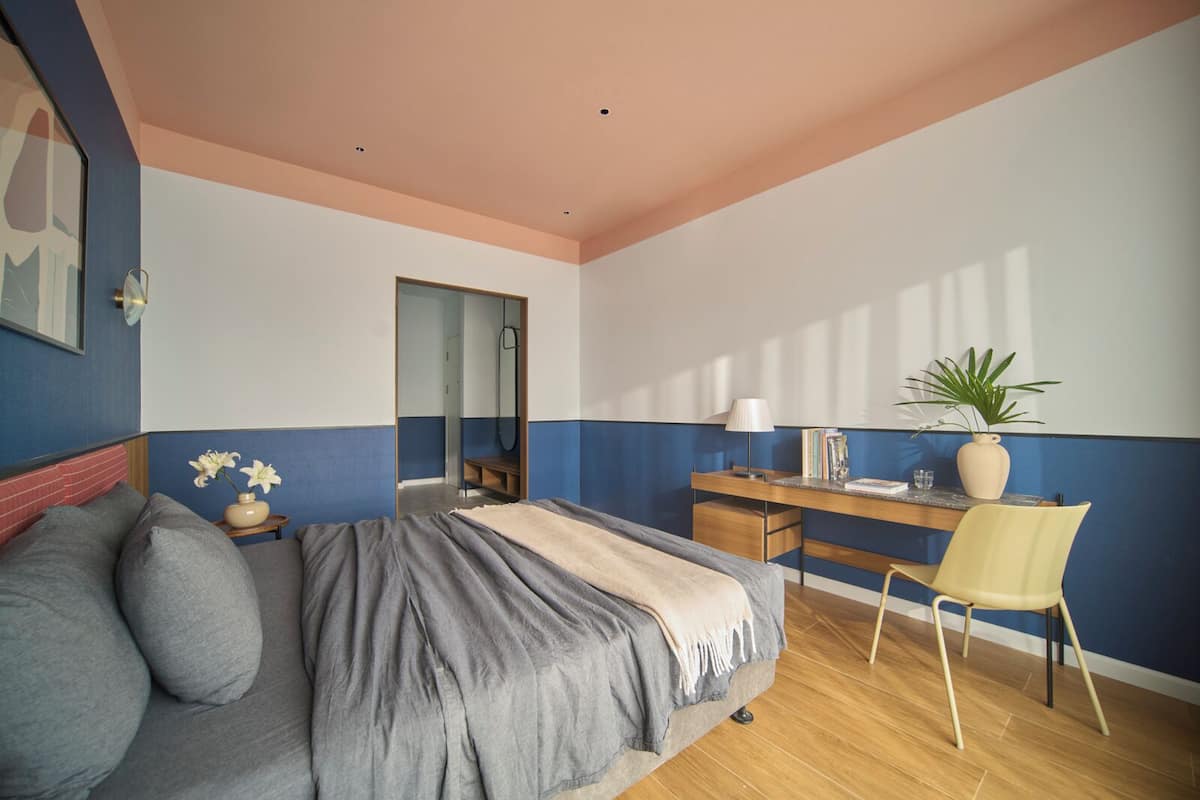 A bedroom with a bed, a desk, a chair, a mirror, and a potted plant.