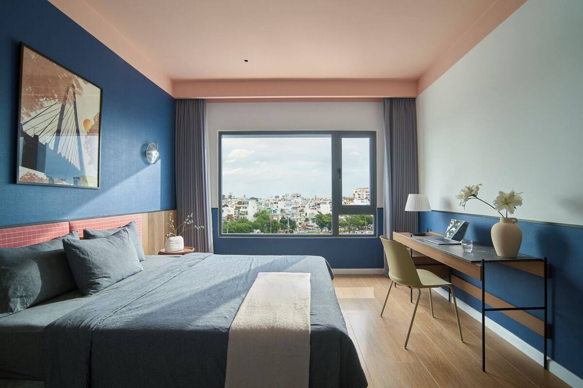A modern hotel room with a large bed, a desk with a laptop, a vase with flowers, and a city view through the window.