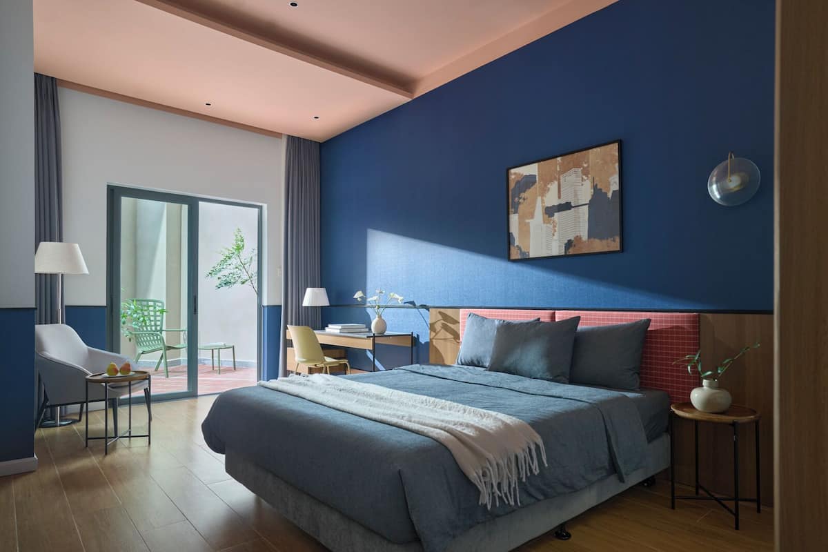 A modern bedroom with a large bed, a desk, and a chair. There is a sliding glass door leading to an outdoor area.
