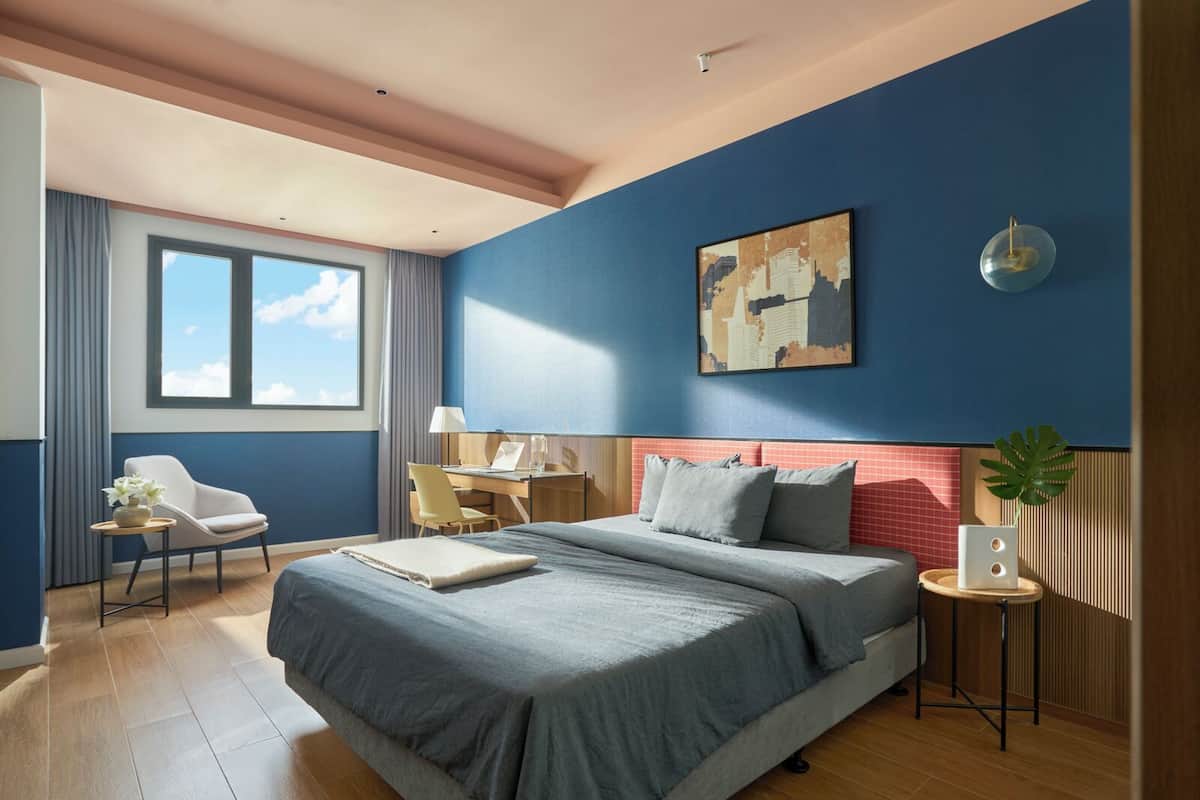 A modern hotel room with a large bed, a desk, a chair, and a window with curtains.