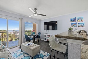 A modern living room with a large window, a ceiling fan, a flat-screen TV, and a dining area with chairs and a table.