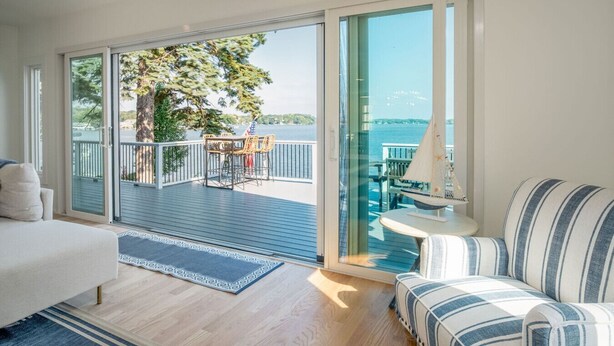A spacious room with a large sliding glass door leading to an outdoor deck with a view of the water and trees. - The “Blue Sapphire” 3BR/3BA Luxury Lakefront Home on Mineola Bay | Stunning Views on Chain O Lakes (Fox Lake)