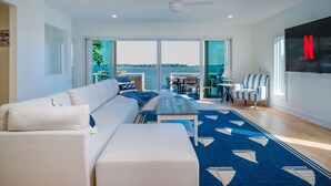 A spacious living room with a large sofa, a coffee table, and a TV mounted on the wall. There is a large window offering a view of the water. - The “Blue Sapphire” 3BR/3BA Luxury Lakefront Home on Mineola Bay | Stunning Views on Chain O Lakes (Fox Lake)