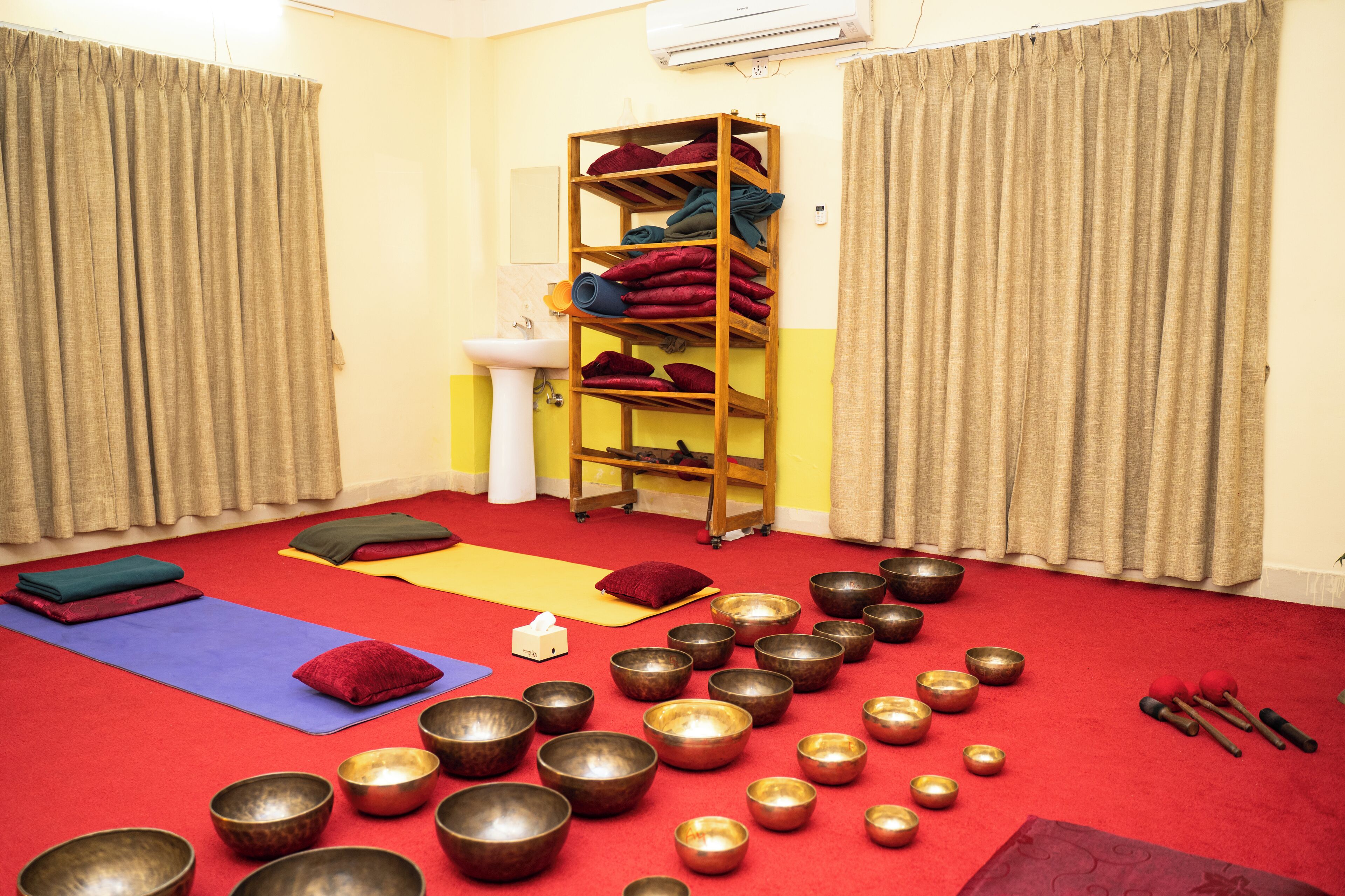 A room with a red carpet, meditation mats, a shelf with cushions, a sink, and multiple bowls.