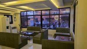 A reception area with a glass partition, cushioned seating, a reception desk, and a view of the street through large windows. - Aarya Wellness & Hostel  (Kathmandu)