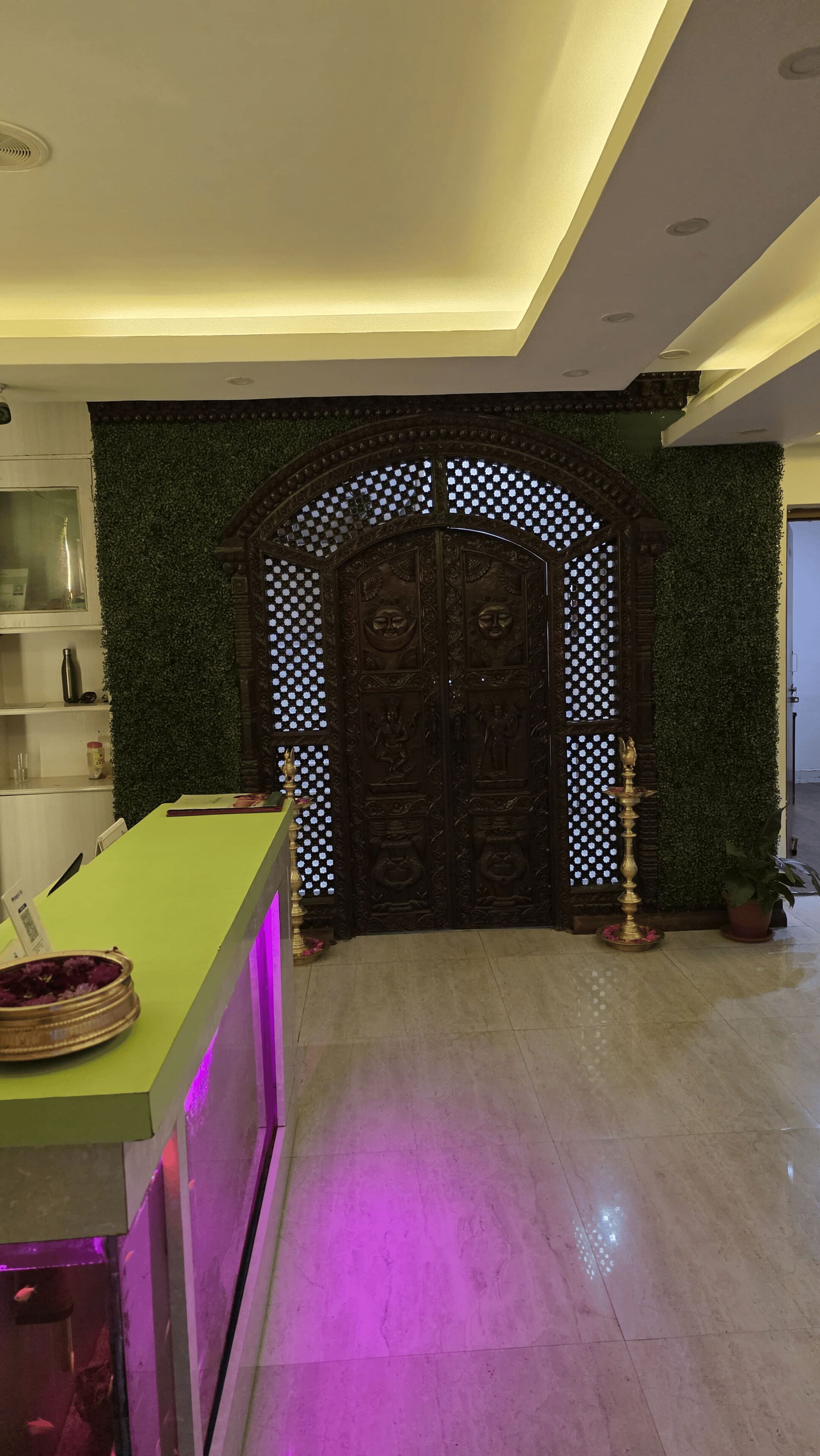 Una zona de recepción con un mostrador verde, una puerta decorativa con detalles intrincados y una pared verde con follaje.
