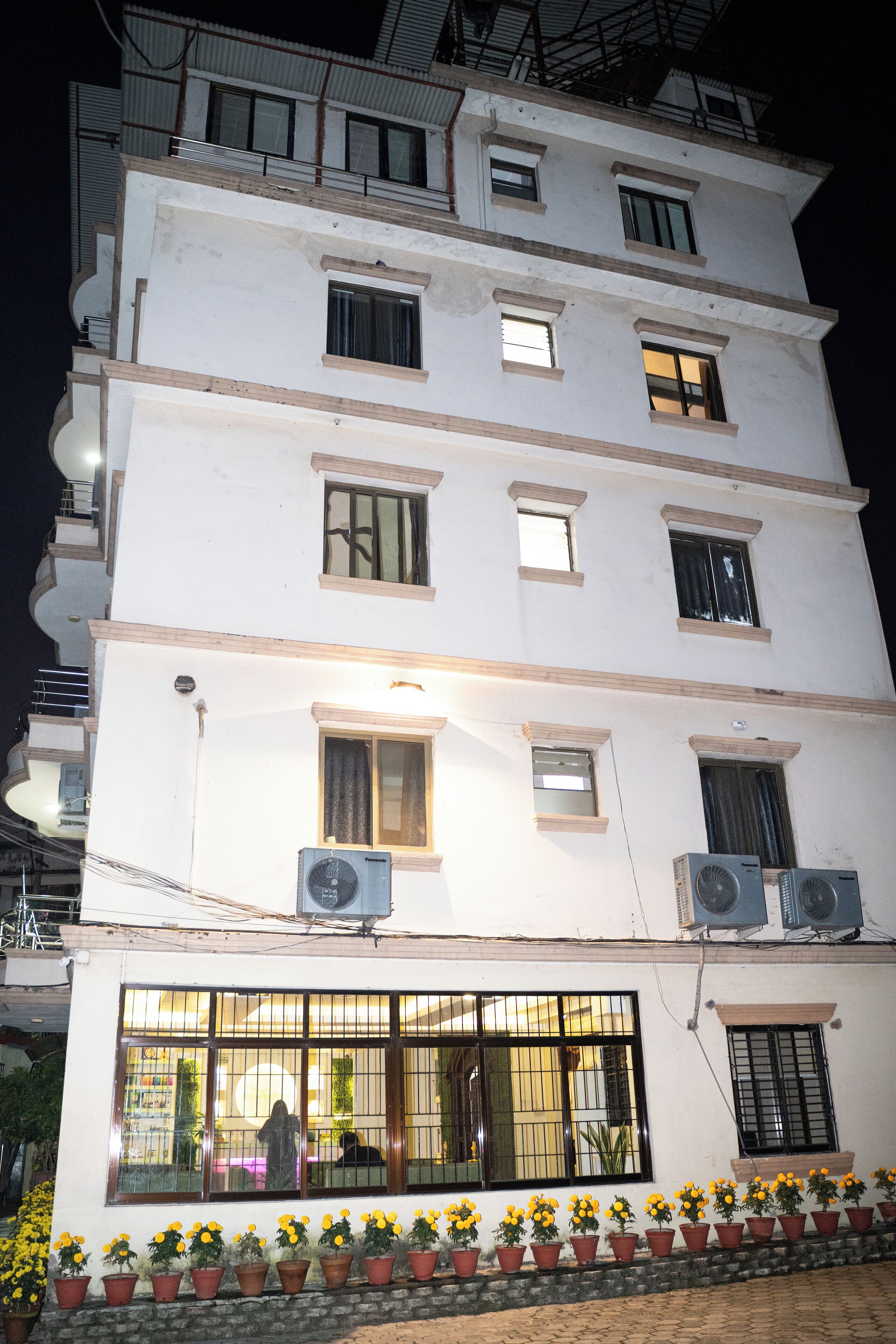 Un edificio de varios pisos con varias unidades de aire acondicionado, un balcón y una fila de plantas en macetas al frente.