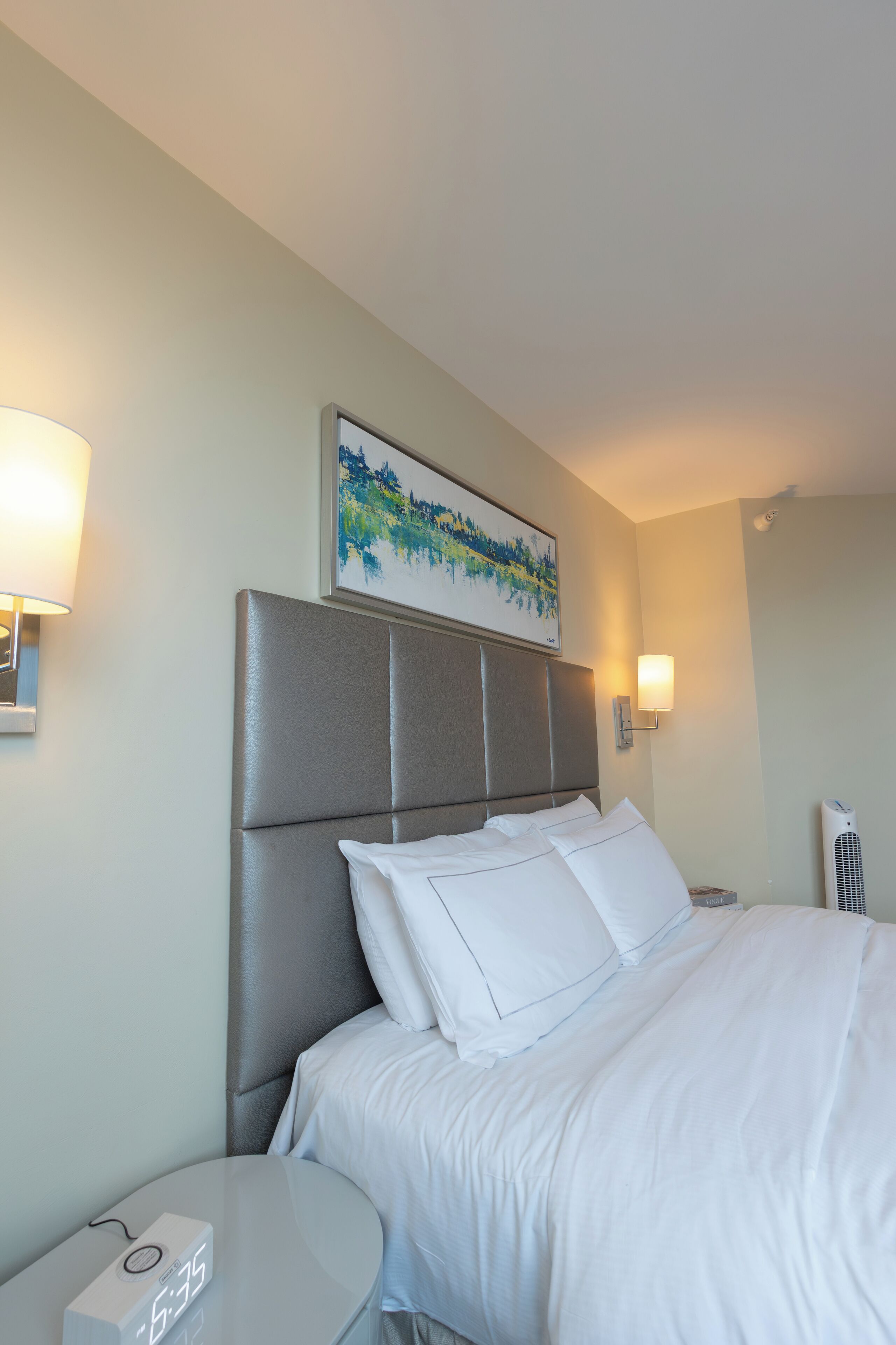 A hotel room with a bed, a bedside table with a clock, and a wall-mounted artwork.