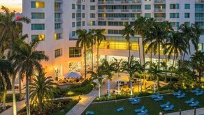 Un hotel de varios pisos con diseño moderno, con balcones en cada planta, un área de piscina exterior bien cuidada con sillones de descanso, y un jardín ajardinado con palmeras.